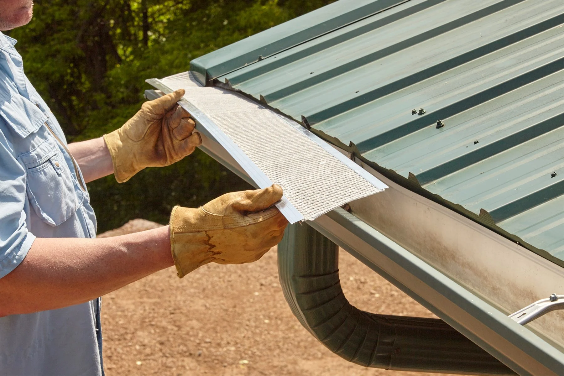 Worker's hand on a roof edge with snow sliding off a shingled roof above a gutter